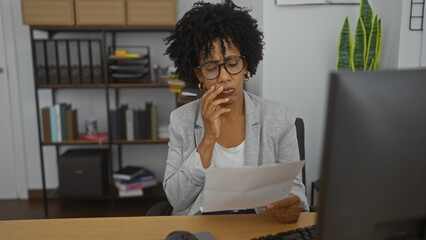 A young african american woman with curly hair is working in an office, reading a document with a...