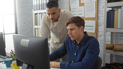 Men working together at a computer in an office environment with shelves and binders in the...