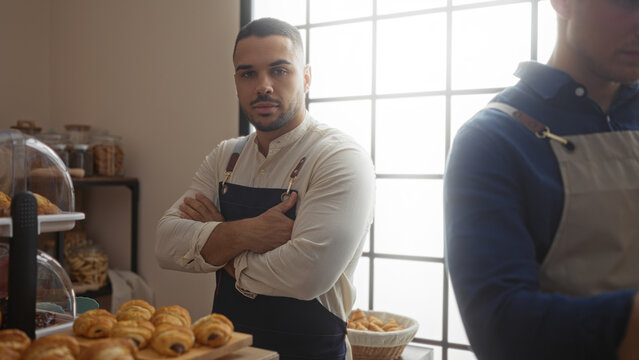 Handsome male bakers working indoors at a bakery shop, displaying various pastries with one man crossing his arms and another focusing on tasks