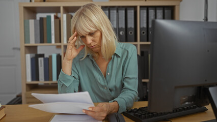 Woman working in an office while looking stressed, middle aged, blonde, reading papers at a desk in an indoor setting.