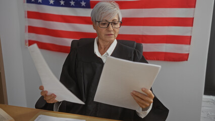 Elderly female judge examines documents in a courtroom with an american flag backdrop, signifying...
