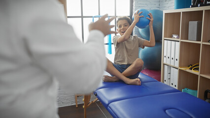 A woman therapist guides a boy with exercise ball during pediatric physiotherapy session in clinic.
