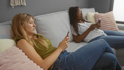 Two women lounging on a sofa in a living room, both focused on their smartphones, creating a relaxed indoor atmosphere.