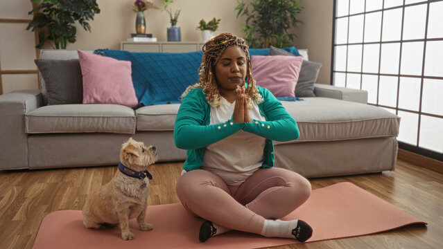 Young african american woman with braids meditating on a yoga mat in her living room with her pet dog.