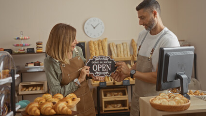 Man and woman workers holding open sign in bakery shop interior with baked goods