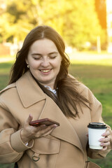 A young woman in a beige coat smiles as she holds a coffee cup and checks her phone in a sunny...