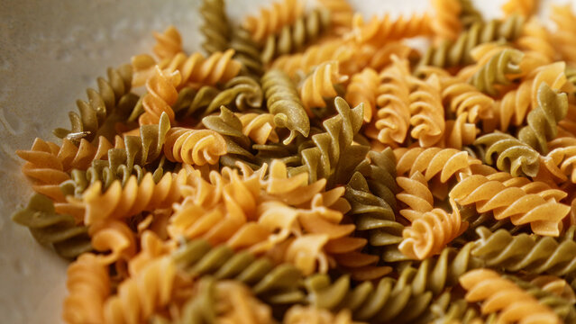 Closeup of uncooked fusilli pasta with green and orange hues arranged in a pile suggesting an italian culinary theme