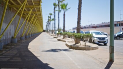 Naklejka premium Defocused street scene featuring palm trees, parked cars, and a modern walkway structure in a sunlit outdoor urban area.
