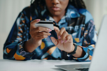 African American woman looking at payment card while using laptop and sitting at table. Focused on task, she wears a colorful shirt and has painted nails