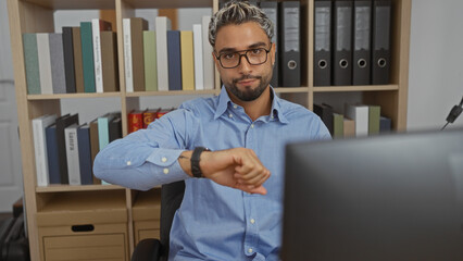 Handsome arab man with beard looking at his watch while sitting at a desk in an office with shelves...