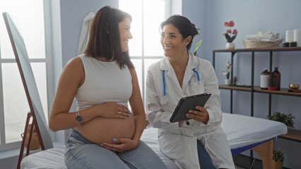 Female doctor using a tablet while consulting a smiling pregnant woman in a clinical room