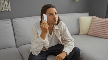 Fototapeta premium Young man with long hair sitting in a living room holding a credit card, appearing thoughtful, indoors in a home setting.