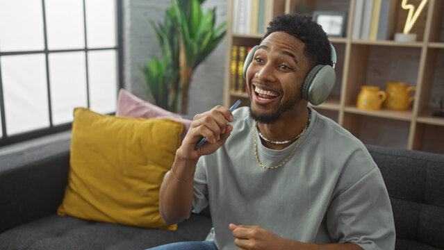 Happy african american man singing with phone as microphone at home on couch with headphones and decor. - Powered by Adobe