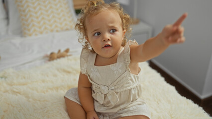 Fototapeta premium Adorable toddler girl with blonde hair pointing while sitting on a bed in a cozy bedroom at home