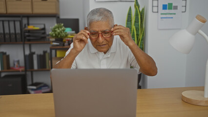 Fototapeta premium Mature hispanic man adjusting glasses while working on a laptop in a well-organized office space with shelves and plants in the background.
