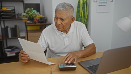 A mature hispanic man with grey hair working in an office, analyzing documents while using a calculator and a laptop, highlighting a focused professional atmosphere.