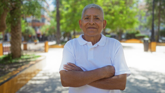 Handsome hispanic grey-haired man standing with crossed arms in an outdoor urban park setting on a sunny day in the city
