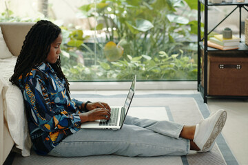 Young African American woman sitting on floor, typing on laptop in bright, modern living space with...