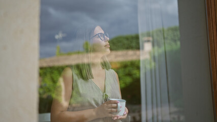 Woman indoors looking through window with reflections holding a cup in quiet cafe setting