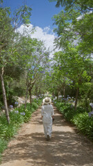 Woman walking along a serene tree-lined path in mallorca, spain, dressed in white with a hat, surrounded by lush greenery and a clear blue sky above.