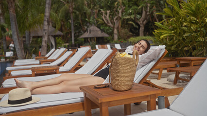 Hispanic woman relaxing at bali resort poolside, vacation tranquility with sunhat and tropical setting.