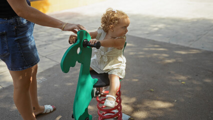 Woman helping a toddler ride a spring rocker at an outdoor playground under the shade of trees in a...
