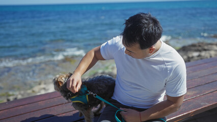 Young asian man sitting with his dog at a seaside promenade in an outdoor city setting with clear blue water in the background.