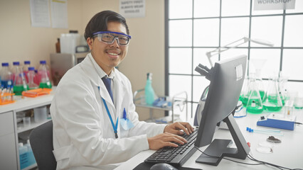 Smiling asian scientist in lab coat working on computer in modern laboratory setting with equipment