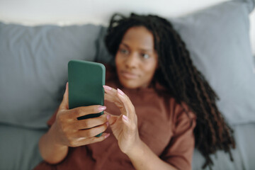 Woman relaxing on bed while engaging with her smartphone, displaying content. Focused and calm, her...