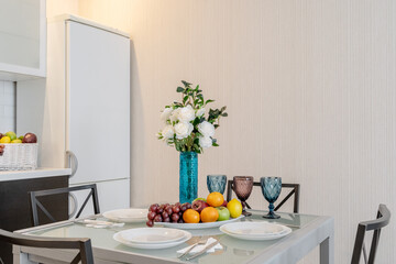 interior design of large modern kitchen. Table setting. On glass table are white plates, colorful glasses, fruits, blue vase with white roses. In background is refrigerator and fruit basket.