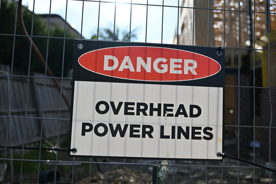 Rectangular danger overhead power lines sign attached to a tilting metal fence, outside the building site of a suburban home