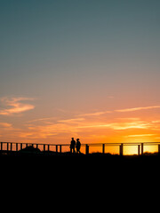 Fototapeta premium Silhouetted couple walks along a boardwalk at sunset, framed by vivid orange and blue skies. A serene, atmospheric moment with ample copy space for text overlay or creative design