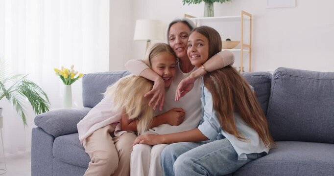 Happy smiling mother sitting on sofa, hugging her two loving preteen daughters and cuddling them up to yourself. Joyful family sharing tender and warm moment of love and affection in their cozy home.