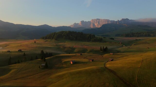 In this drone footage, the first light of dawn spreads over the rolling fields and wooden farmhouses of Seiser Alm, casting shadows across the landscape framed by the Dolomite mountains.