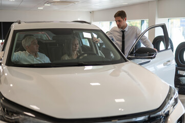 A gray-haired Caucasian woman is sitting behind the wheel of a car. Car dealership salesman consulting mature married couple.