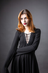 Red-haired gothic girl in a black dress on a gloomy background in the studio.