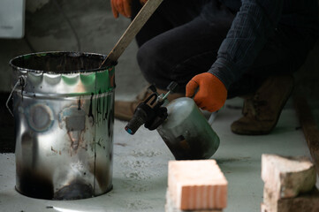 A man uses a kerosene lamp to heat resin in a bucket, working with waterproofing and resin.