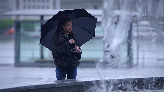 A person holding an umbrella stands alone in a rainy, urban environment filled with reflection