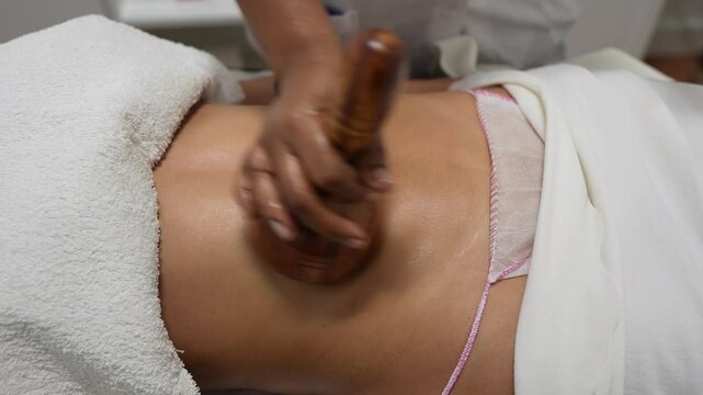 Young woman is lying on a massage table and smiling while receiving maderotherapy treatment on her stomach