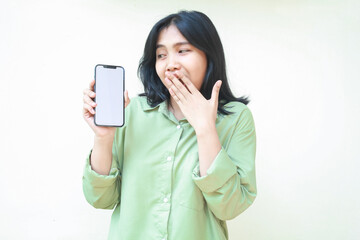 surprised dark hair asian woman looking aside showing smartphone white screen and covering her open mouth with hands wearing green oversize shirt isolated on white background