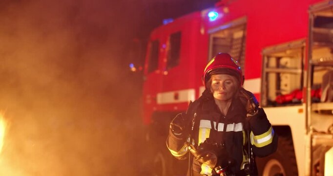 Caucasian young woman firefighter coming out from fire and smoke outside and taking out helmet. Female hero fireguard at red water truck. Hard breathing. Tired firewoman go resting.