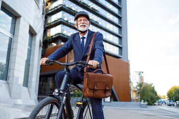 Low angle view of smiling old businessman with shoulder bag and helmet riding bicycle to work in...