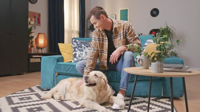 Young man playing with his golden retriever pet on living room floor in modern apartment. Support, friendship, love and care. Handsome happy guy relaxing on couch, playing with his loyal puppy.