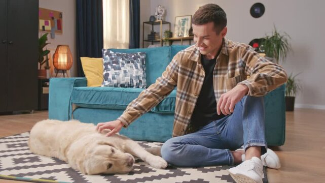 Male pet owner sitting at floor with golden retriever dog in home bedroom. Handsome young man spending time with sleeping pet, petting him, bonding. Calming and relaxing. Moment of togetherness.