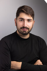 Fototapeta premium portrait d'un beau jeune homme de 25 ans. Il est brun, barbu et souriant et il porte un pull over noir. Arrière plan gris.