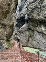 A narrow pathway winds through Partnach Gorge beside a clear blue, rushing stream, with red leaves scattered on the ground, creating a dramatic autumn landscape in Bavaria.