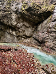 Wet rocks line the vibrant turquoise stream flowing through Partnach Gorge, with red leaves scattered along the ground, creating a striking autumn contrast in the Bavarian Alps.