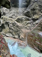 A path winds through the rocky cliffs of Partnachklamm Gorge, with a stream at its base and misty droplets from nearby waterfalls creating an enchanting atmosphere, Garmisch-Partenkirchen.