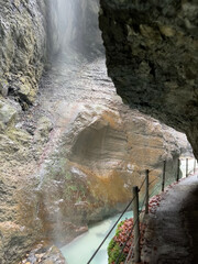 A path winds through the rocky cliffs of Partnachklamm Gorge, with a stream at its base and misty droplets from nearby waterfalls creating an enchanting atmosphere, Garmisch-Partenkirchen.