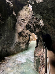 A narrow walkway winds through the rock cliffs at the valley's base alongside a flowing stream at Partnachklamm Gorge, Garmisch-Partenkirchen, offering a stunning alpine experience.